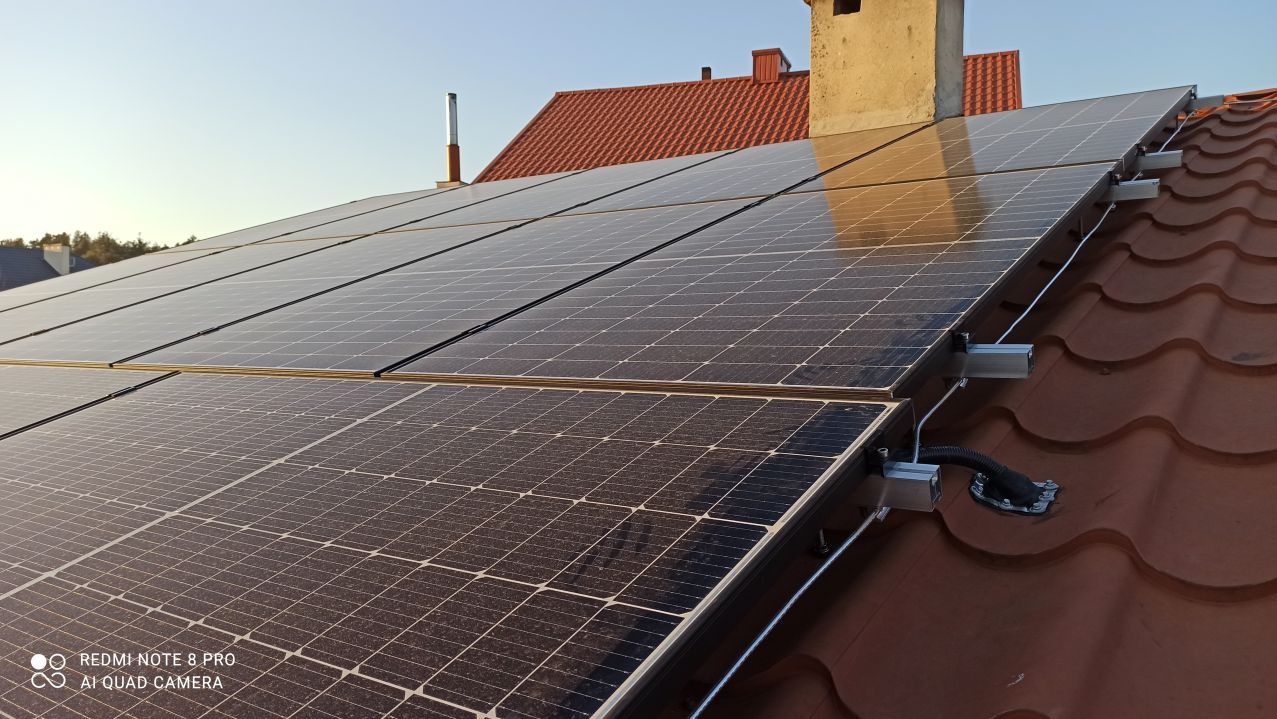 Panele słoneczne zamontowane na dachu z blachodachówki w promieniach popołudniowego słońca, widoczne kable i elementy montażowe.