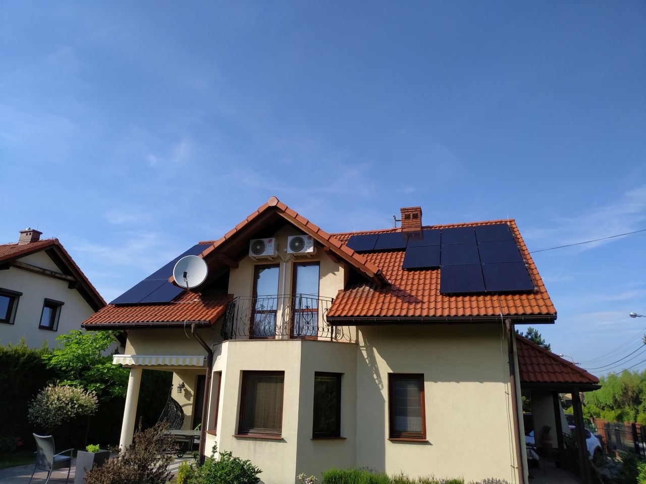 Dom jednorodzinny z panelami słonecznymi na dachu, anteną satelitarną i dwoma klimatyzatorami na elewacji, w słoneczny dzień.