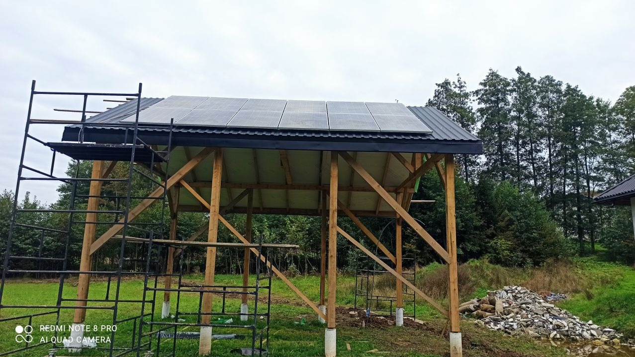 Montaż paneli na drewnianej konstrukcji z metalowym dachem, widoczne rusztowanie i przygotowany teren.