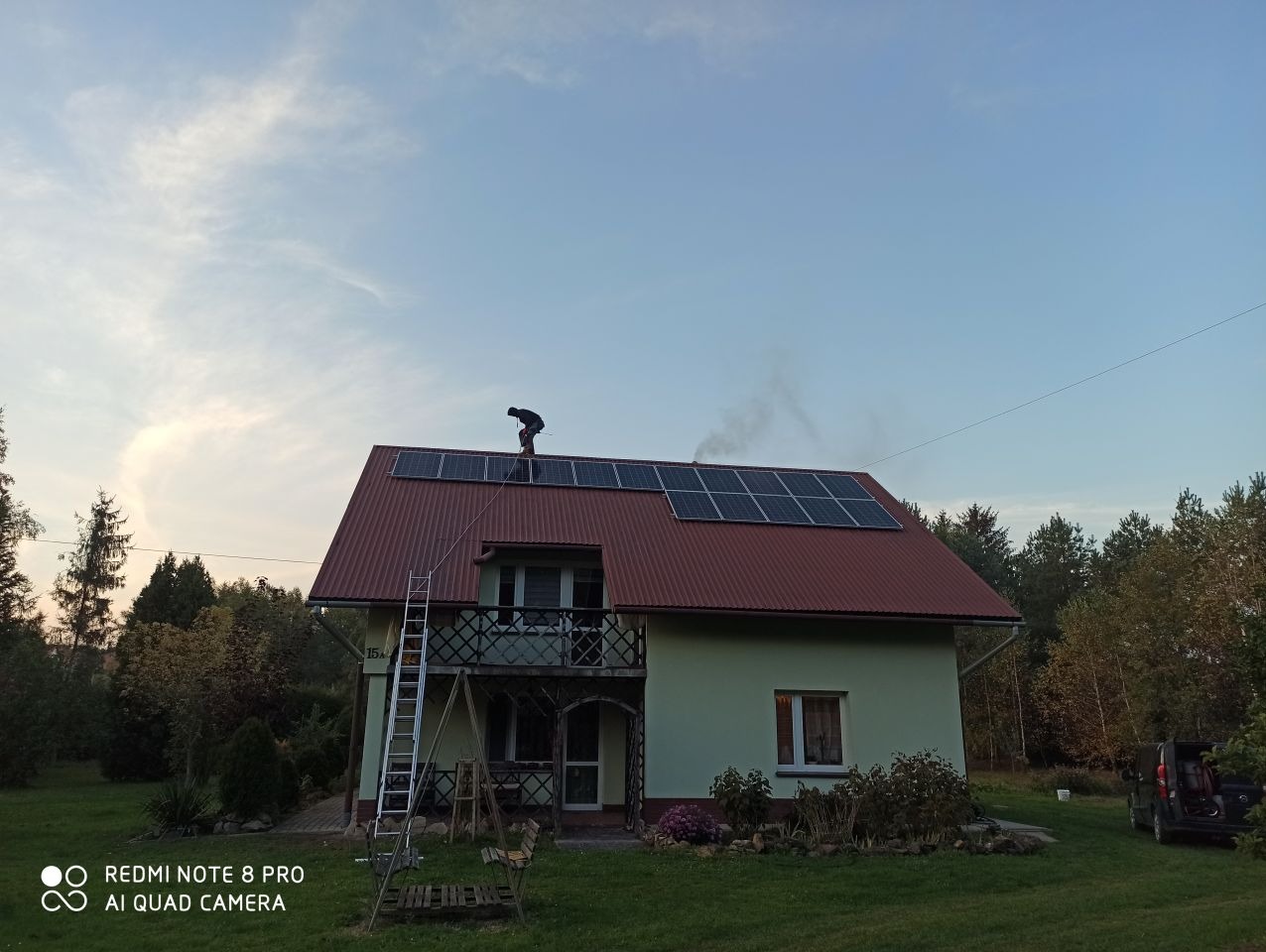 Montaż paneli słonecznych na dachu dwukondygnacyjnego domu jednorodzinnego z drabiny, widok z dołu na tle zachmurzonego nieba i zieleni drzew.