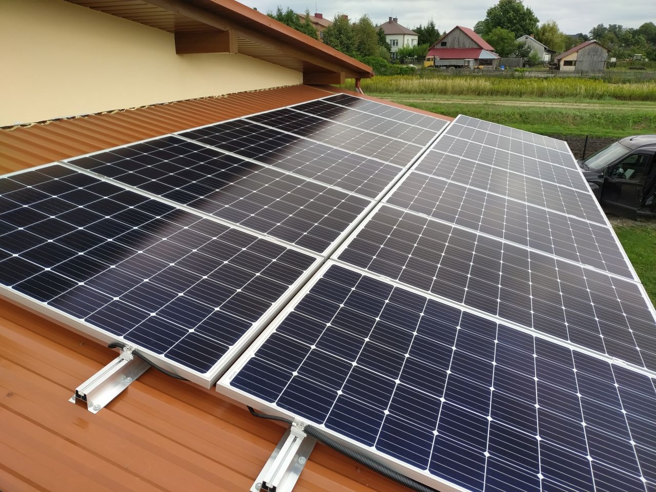 Panele słoneczne zamontowane na brązowym dachu blaszanym, widok z góry, w tle domy jednorodzinne i zielone pola.