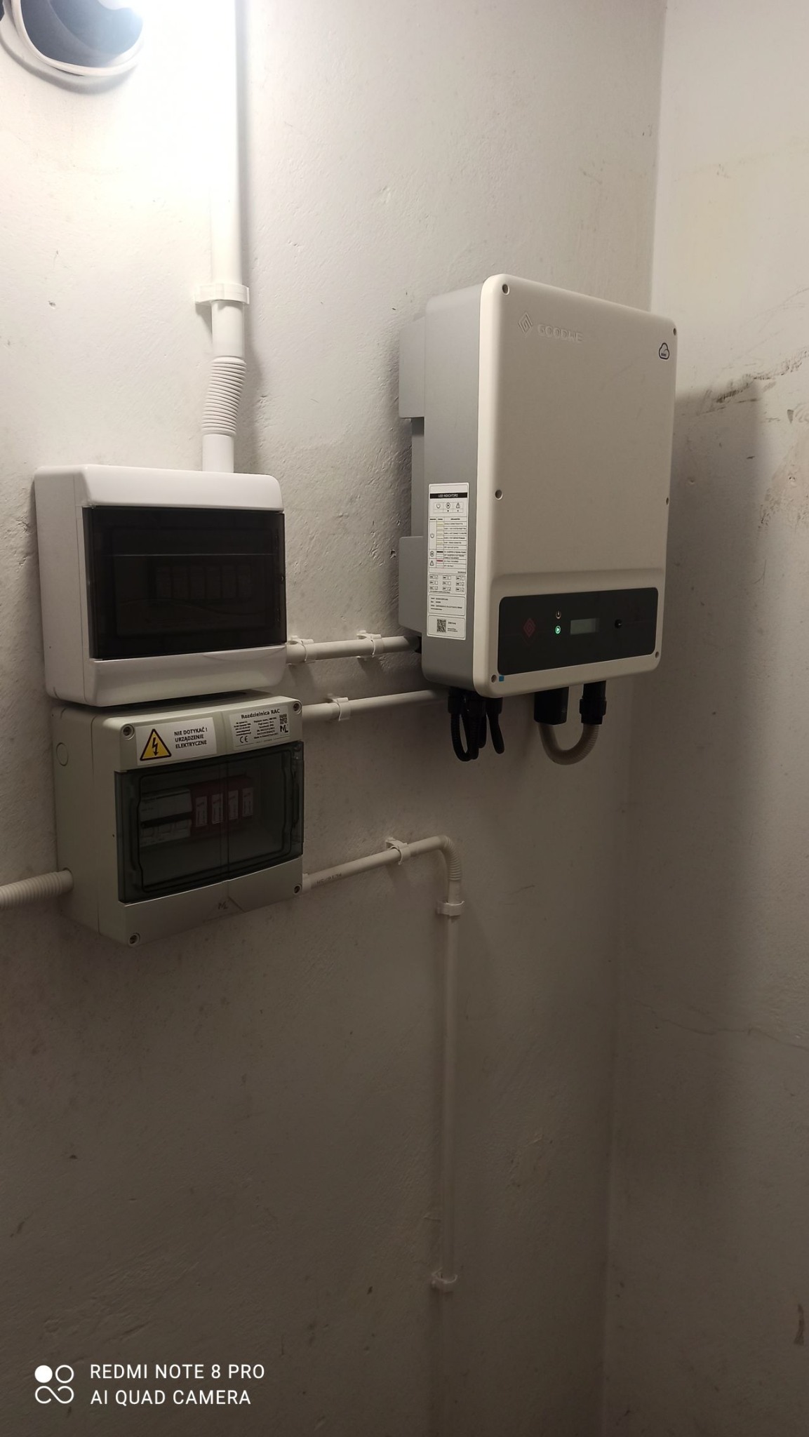 Szare skrzynki elektryczne i inwerter Goodwe zamontowane na pomalowanej ścianie wewnątrz budynku, połączone białymi rurami PCV.