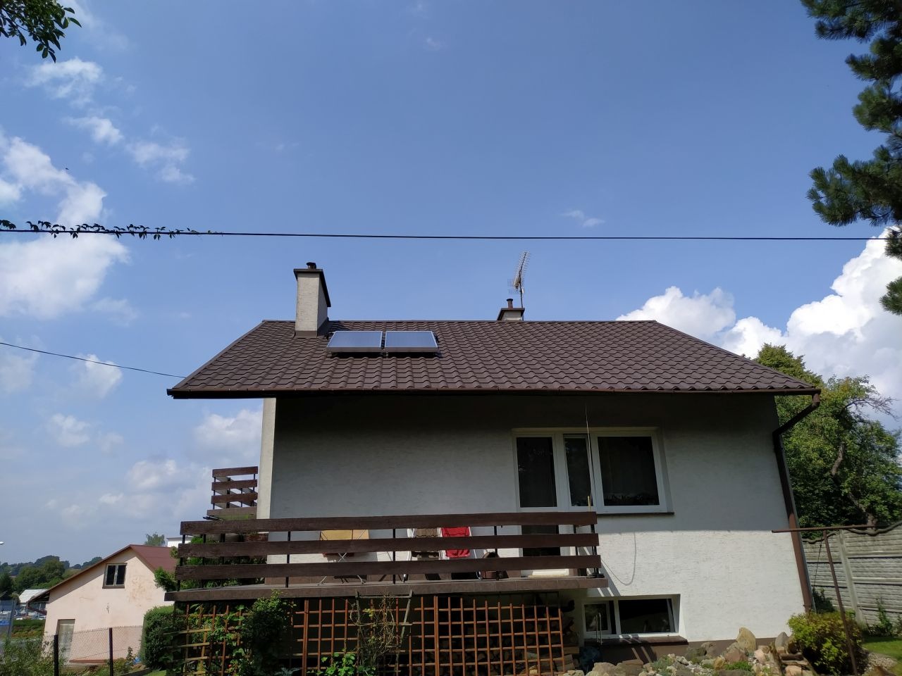 Dom jednorodzinny z brązowym dachem, na którym zamontowane są dwa płaskie, prostokątne kolektory słoneczne. Widoczny komin, antena oraz balkon z drewnianą balustradą. Na pierwszym planie ogródek...