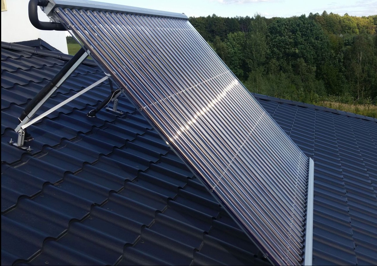 Rząd szklanych rur kolektora słonecznego na ciemnym dachu z metalowej dachówki, wsparty na białej metalowej konstrukcji, z widocznymi czarnymi rurami izolacyjnymi, na tle zieleni drzew.
