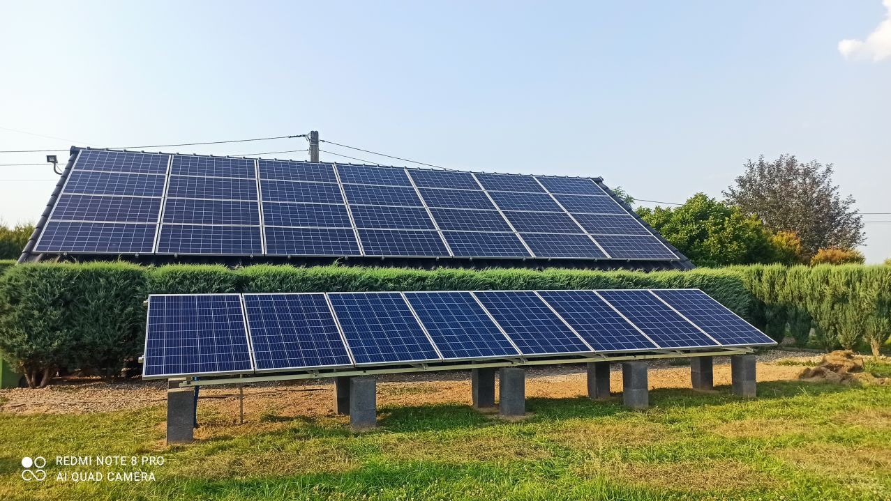 Instalacja paneli słonecznych w Iwierzycach: panele na dachu domu i dodatkowy rząd paneli na betonowych podstawach, otoczony zielenią.