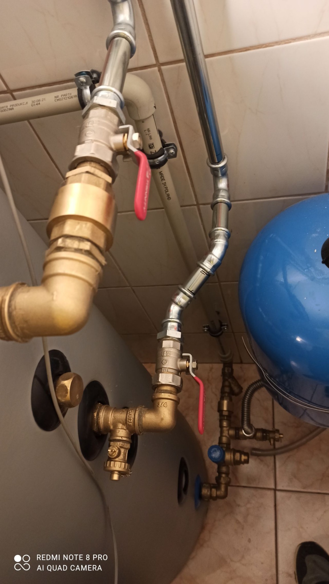 Szczegółowe ujęcie instalacji hydraulicznej z rurami miedzianymi i zaworami z czerwonymi uchwytami, połączonymi z szarym zasobnikiem i niebieskim zbiornikiem wyrównawczym, widoczne płytki...