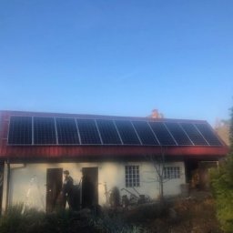 P.H.U.MEGALUX SP.Z O.O. - Dom jednorodzinny z panelami słonecznymi na czerwonym dachu, drabina oparta o ścianę, instalator na dachu, widoczne drzewa w tle.