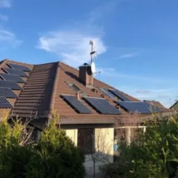 Panele słoneczne zamontowane na brązowym dachu domu w Policach, widoczne z poziomu ogrodu, antena satelitarna na kominie.