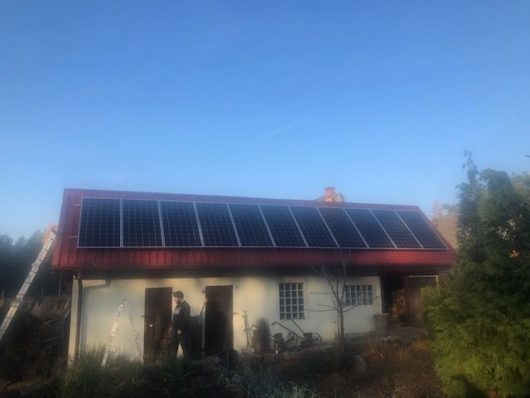 Dom jednorodzinny z panelami słonecznymi na czerwonym dachu, drabina oparta o ścianę, instalator na dachu, widoczne drzewa w tle.