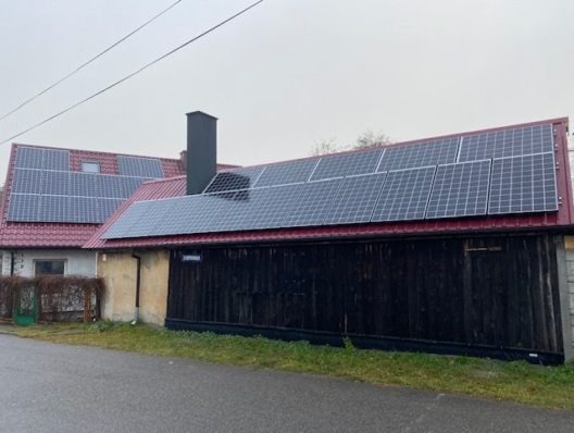 Instalacja fotowoltaiczna na dachu budynku z czerwoną dachówką, widok z ulicy w pochmurny dzień.