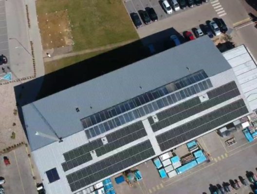 Widok z drona na duży budynek przemysłowy z panelami słonecznymi na dachu, otoczony parkingiem i placem składowym.