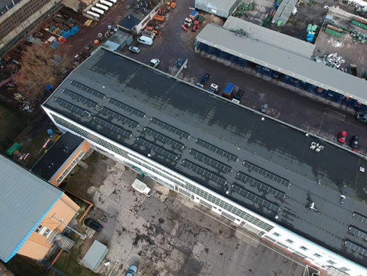 Widok z lotu ptaka na duży budynek z panelami słonecznymi na dachu, otoczony innymi budynkami i placem manewrowym.