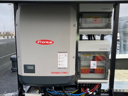 Szary inwerter Fronius Symo z widocznymi skrzynkami zabezpieczeń na dachu budynku, okablowanie w peszlach.