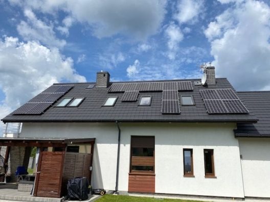 Dom jednorodzinny z panelami słonecznymi zamontowanymi na dachu pokrytym ciemną dachówką, widoczne okna dachowe i komin z anteną satelitarną, błękitne niebo z chmurami w tle.