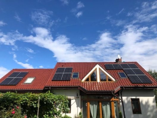 Dom jednorodzinny z czerwoną dachówką i panelami słonecznymi na dachu, błękitne niebo z chmurami w tle.