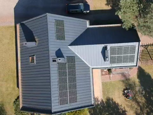 Widok z góry na dom z szarym dachem, na którym zamontowano panele słoneczne. Na podjeździe stoi ciemny samochód, a w ogrodzie widać kosiarkę.