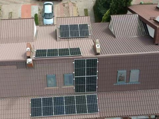 Widok z góry na brązowy dach z zamontowanymi panelami fotowoltaicznymi różnej wielkości i konfiguracji, z kominem i oknami dachowymi, oraz zaparkowanym białym samochodem w tle.