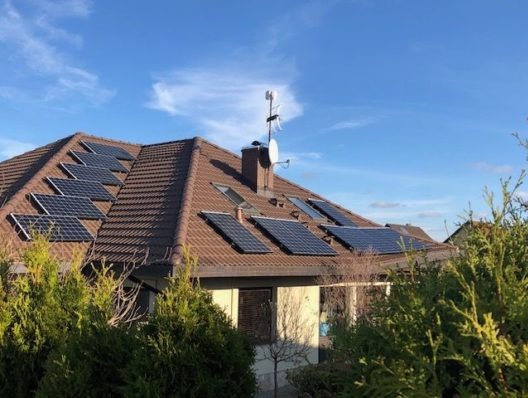 Panele słoneczne zamontowane na brązowym dachu domu w Policach, widoczne z poziomu ogrodu, antena satelitarna na kominie.