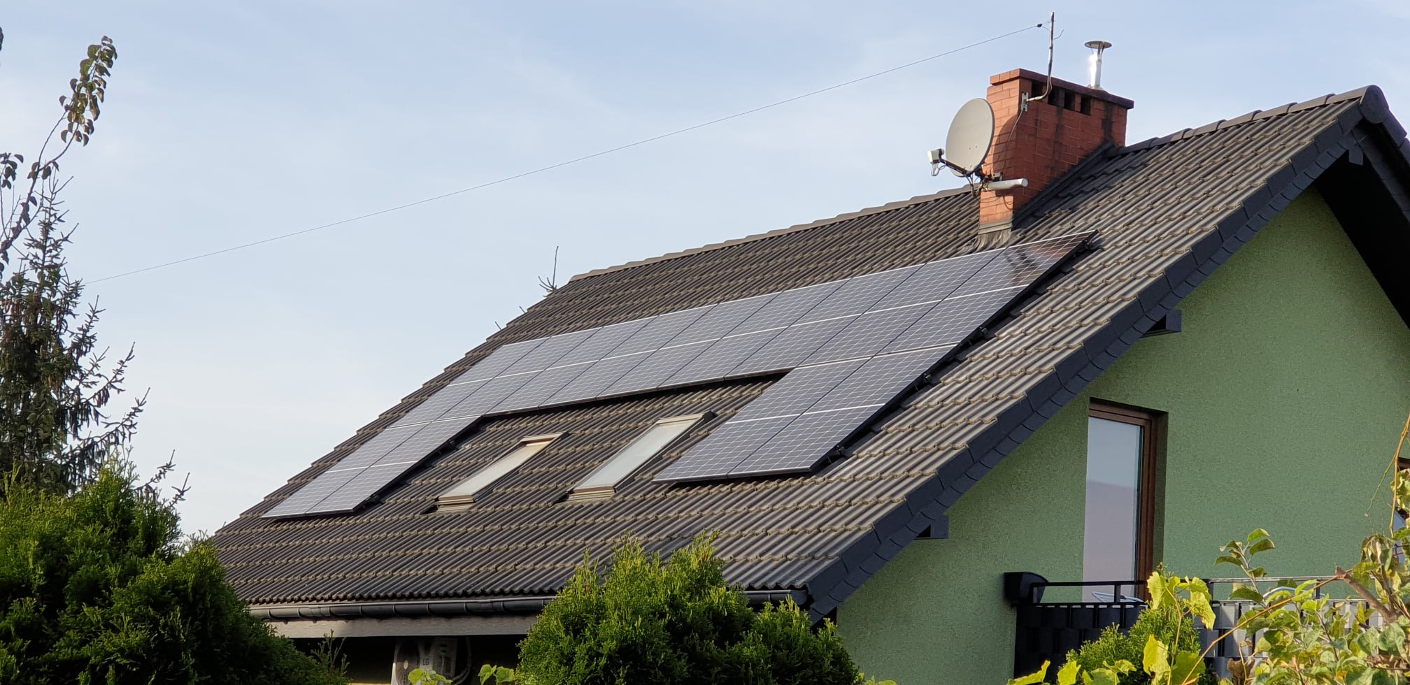 Panele fotowoltaiczne na dachu domu jednorodzinnego z dachówką bitumiczną, widoczny komin z anteną satelitarną i okno dachowe.