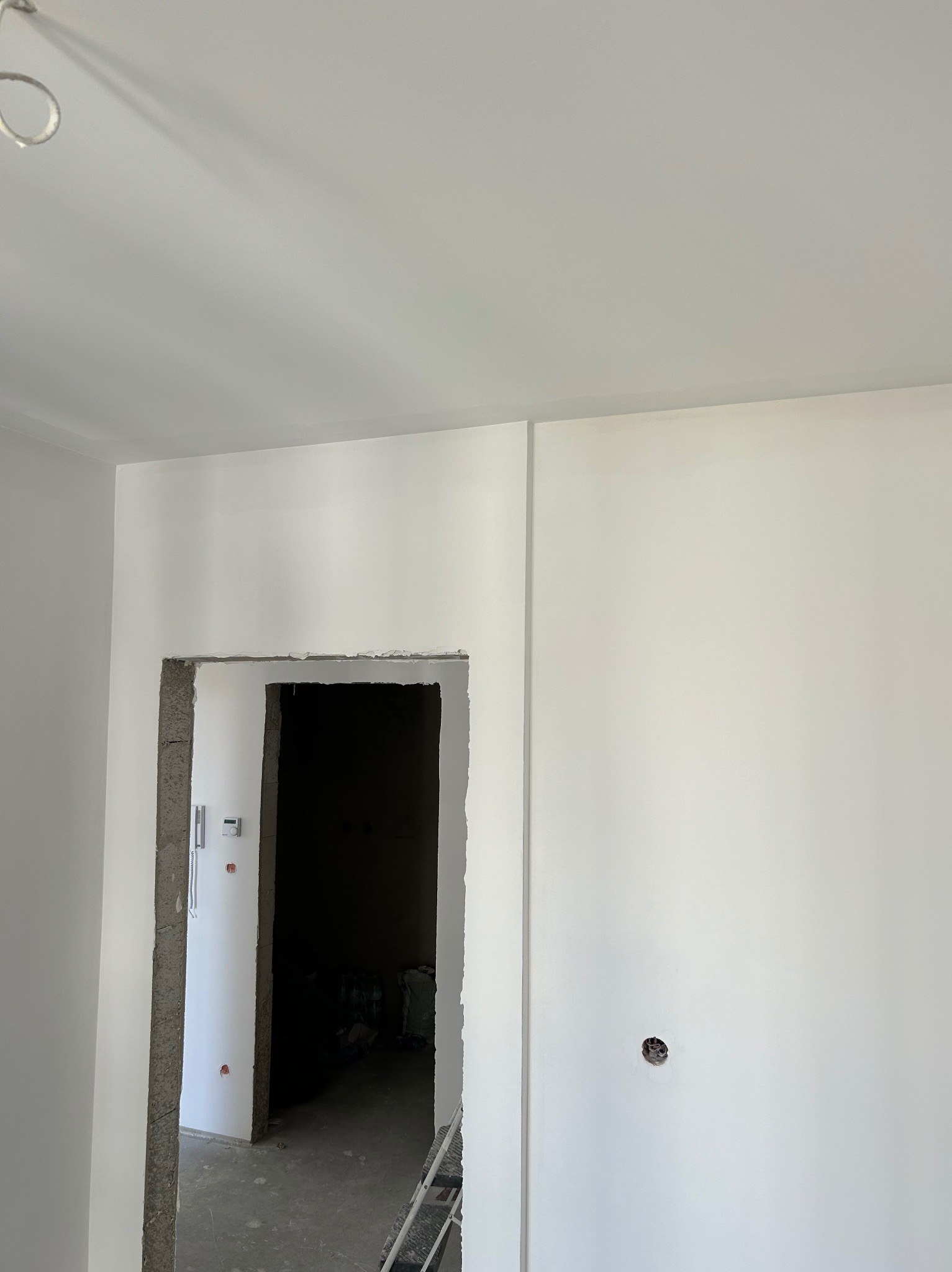 Świeżo pomalowane na biało wnętrze z widocznym otworem drzwiowym w stanie surowym, z wystającymi kablami i widoczną instalacją elektryczną, na podłodze widoczne ślady po remoncie.