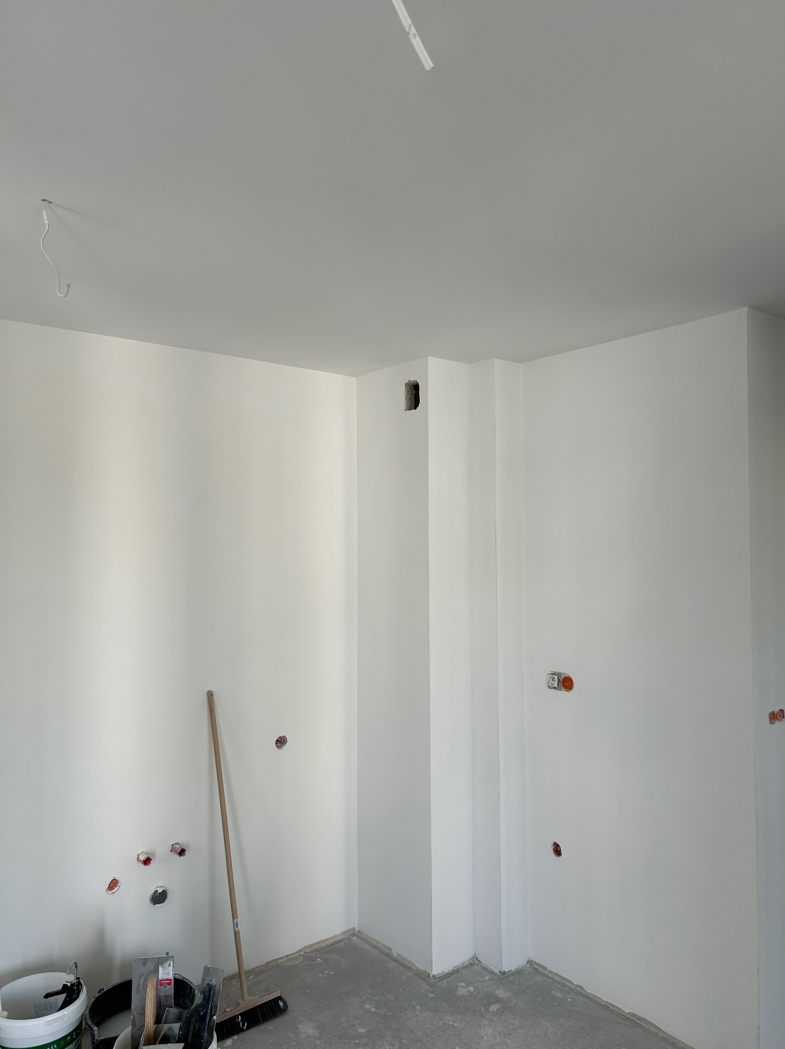 Świeżo pomalowane na biało wnętrze z widocznymi puszkami elektrycznymi, narzędziami malarskimi w wiadrach i miotłą opartą o ścianę, surowy beton na podłodze.