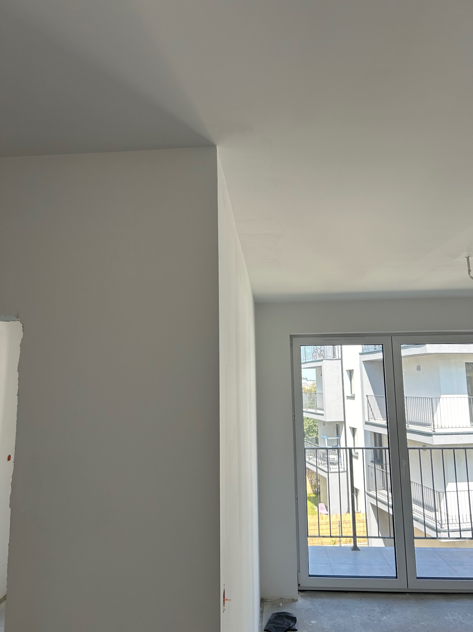 Świeżo pomalowane na biało ściany i sufit w nowym mieszkaniu z widokiem na balkon innego budynku przez duże okno balkonowe.