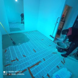 Paweł Michalski - Przygotowanie podłoża pod wylewkę: siatka grzewcza zamocowana taśmą, pracownik miesza zaprawę w wiadrze używając mieszadła elektrycznego, w tle widoczne narzędzia i odzwierciedlenie w lustrze.