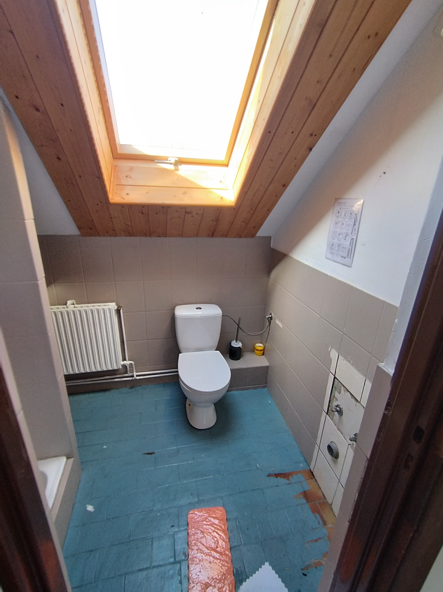 Łazienka na poddaszu ze skośnym sufitem i oknem dachowym, widoczne płytki na ścianach i podłodze w trakcie remontu, z fragmentami starej glazury odsłoniętymi przy podłodze.