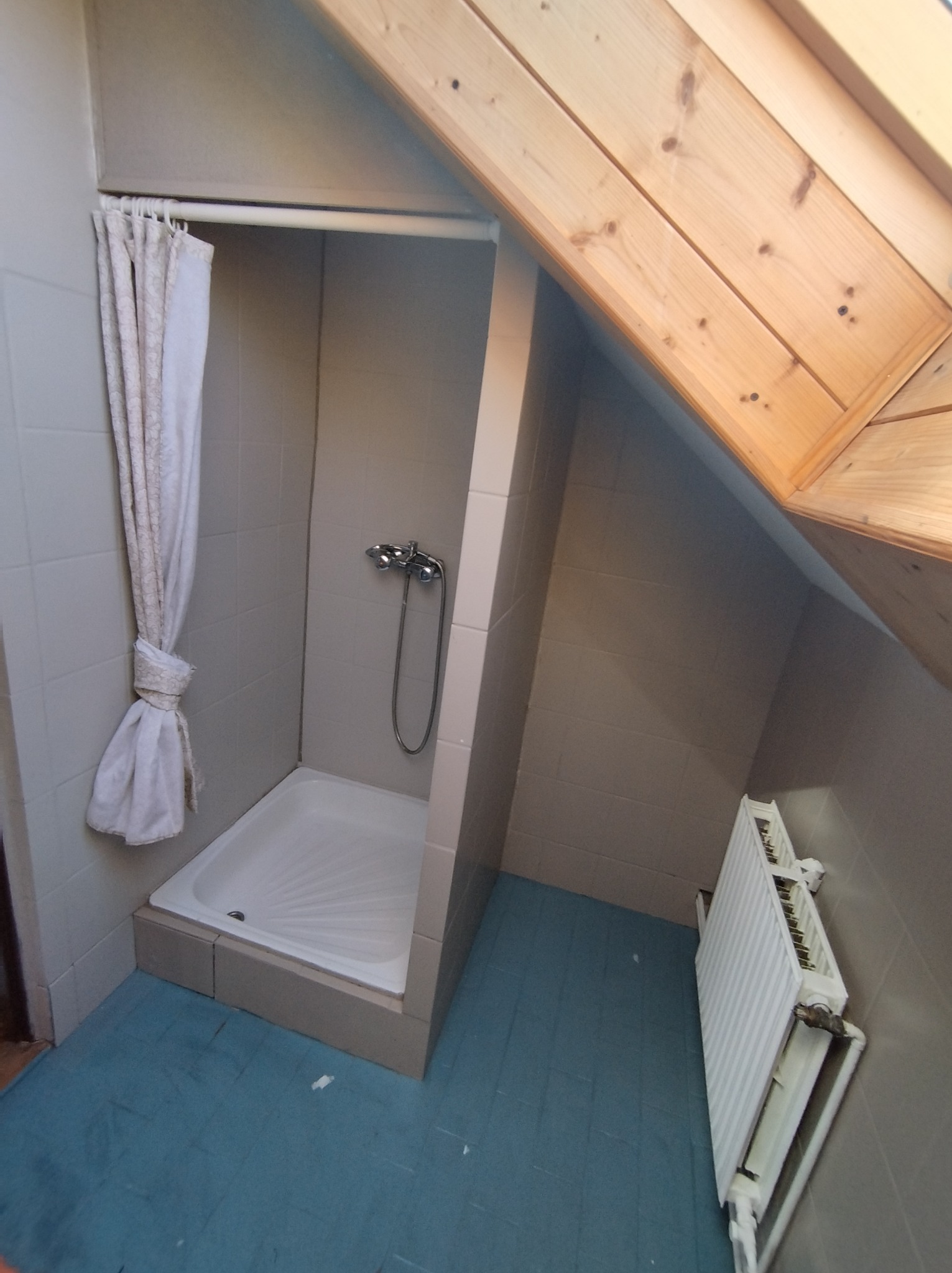 Niewielka kabina prysznicowa z brodzikiem, zasłonięta częściowo kremową zasłoną, umieszczona w łazience na poddaszu z drewnianym wykończeniem dachu i grzejnikiem na ścianie.