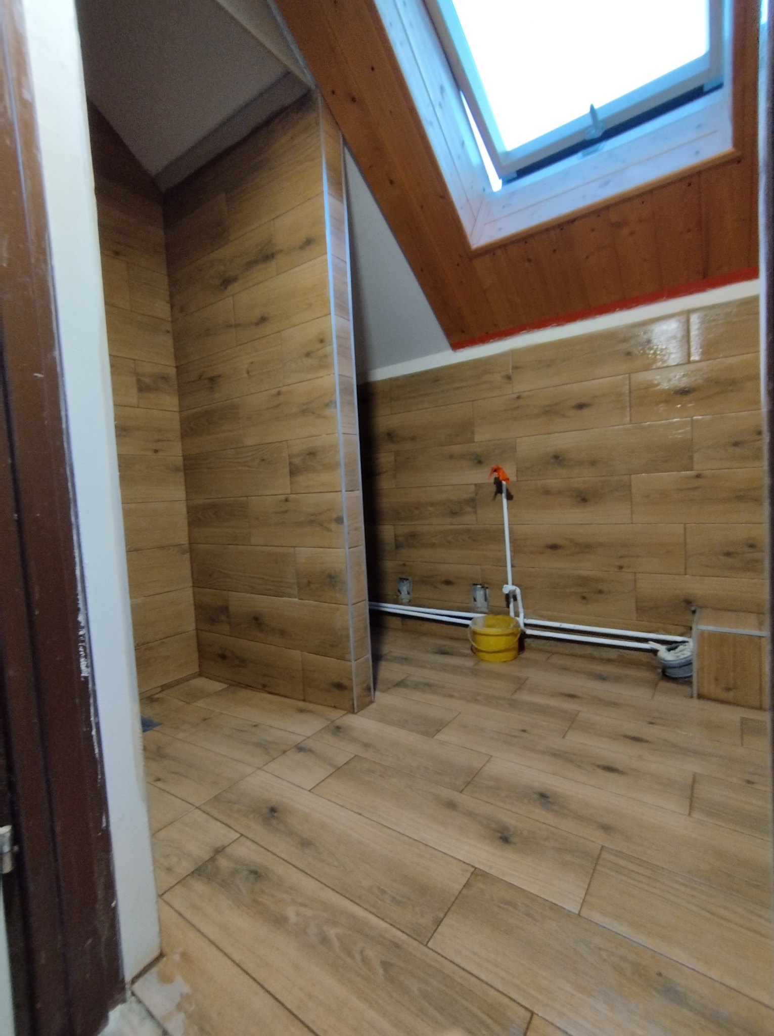 Ukończona łazienka na poddaszu w Koszalinie z drewnopodobnymi płytkami na podłodze i ścianach, widoczne rury i żółte wiadro, oświetlona przez okno dachowe.