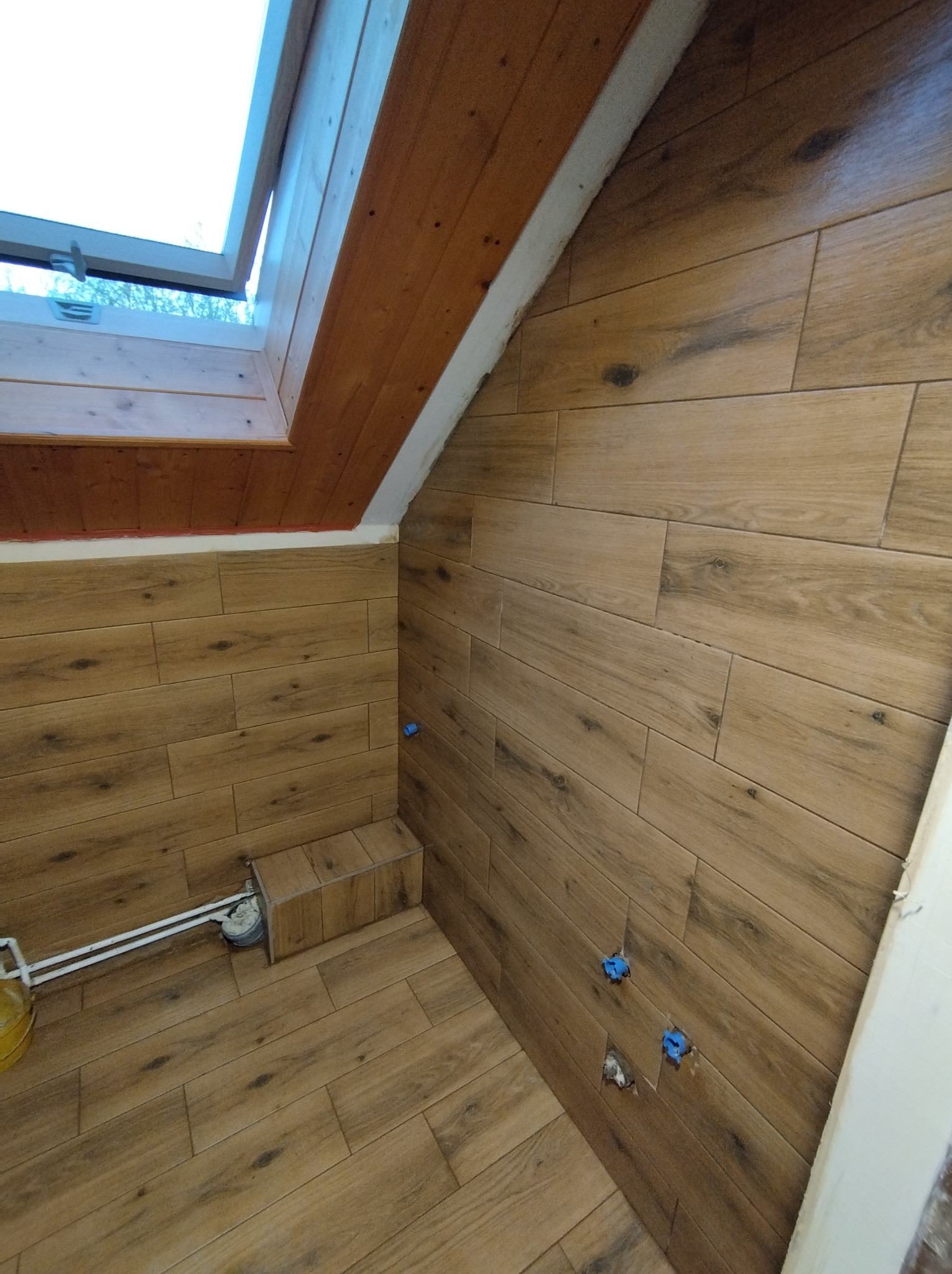 Łazienka na poddaszu z oknem dachowym, wykończona płytkami imitującymi drewno, widoczne niedokończone instalacje i fragment podłączenia do kanalizacji.