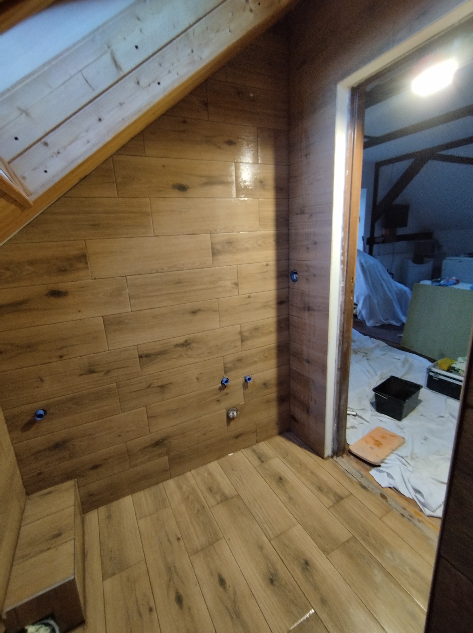 Wykończona łazienka na poddaszu z drewnopodobnymi płytkami na ścianach i podłodze, widoczne wystające rury instalacyjne i akcesoria glazurnicze w sąsiednim pomieszczeniu.