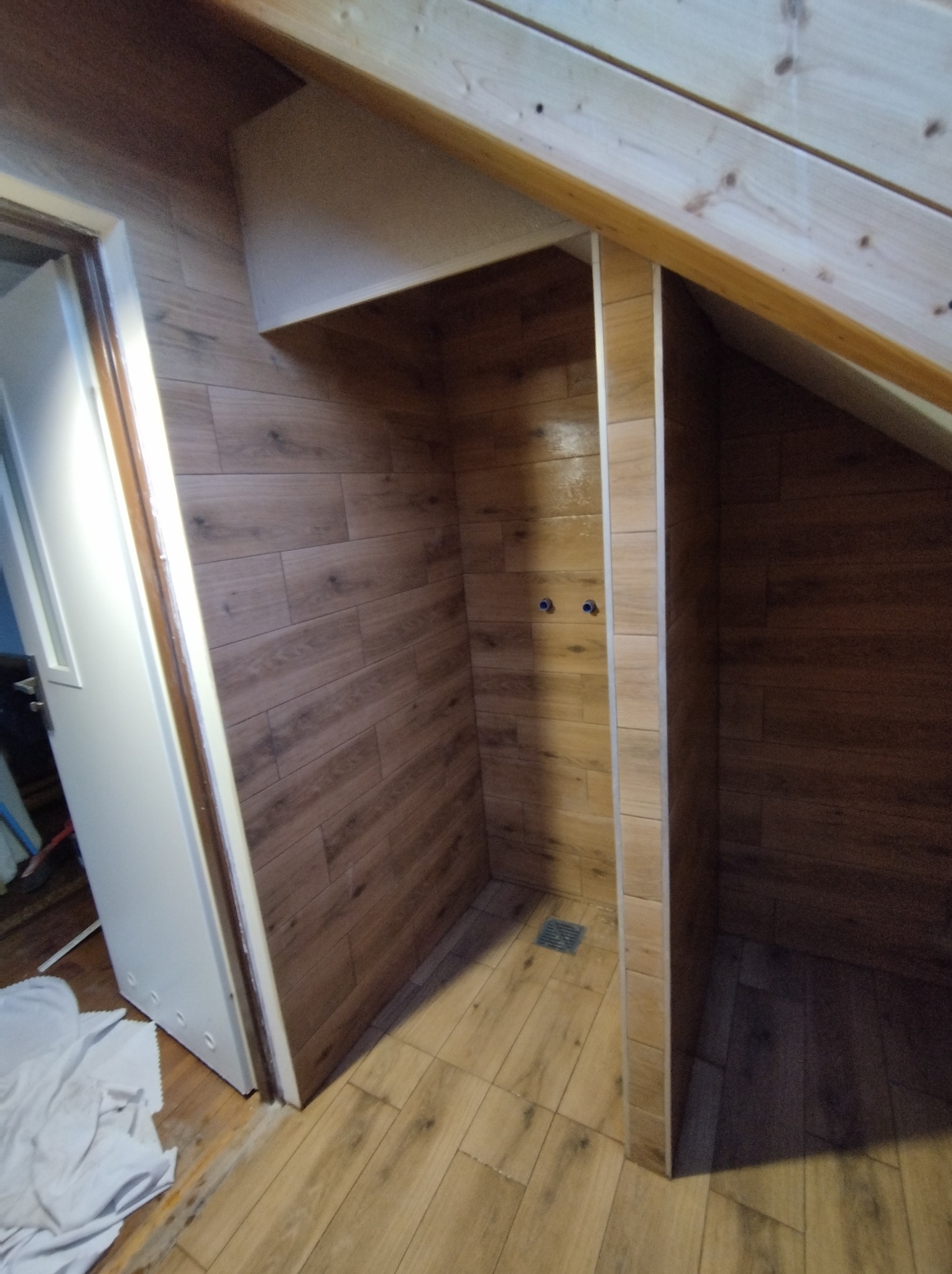 Niewielka kabina prysznicowa w skosie poddasza, wykończona płytkami imitującymi drewno, z widocznym odpływem i surowymi końcówkami rur.