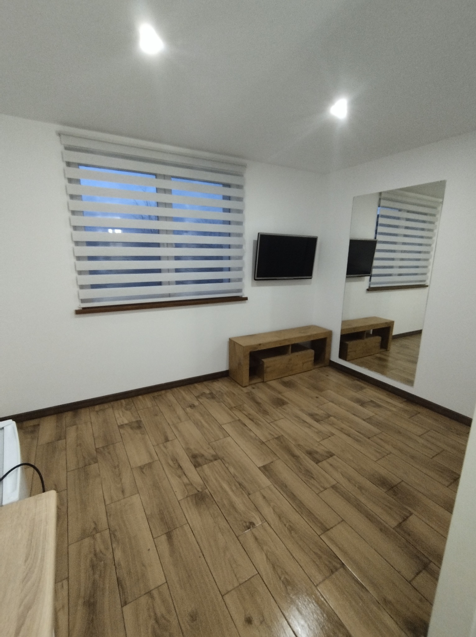 Salon z drewnopodobną podłogą, roletami w oknie i dużym lustrem na ścianie, odbijającym pokój.