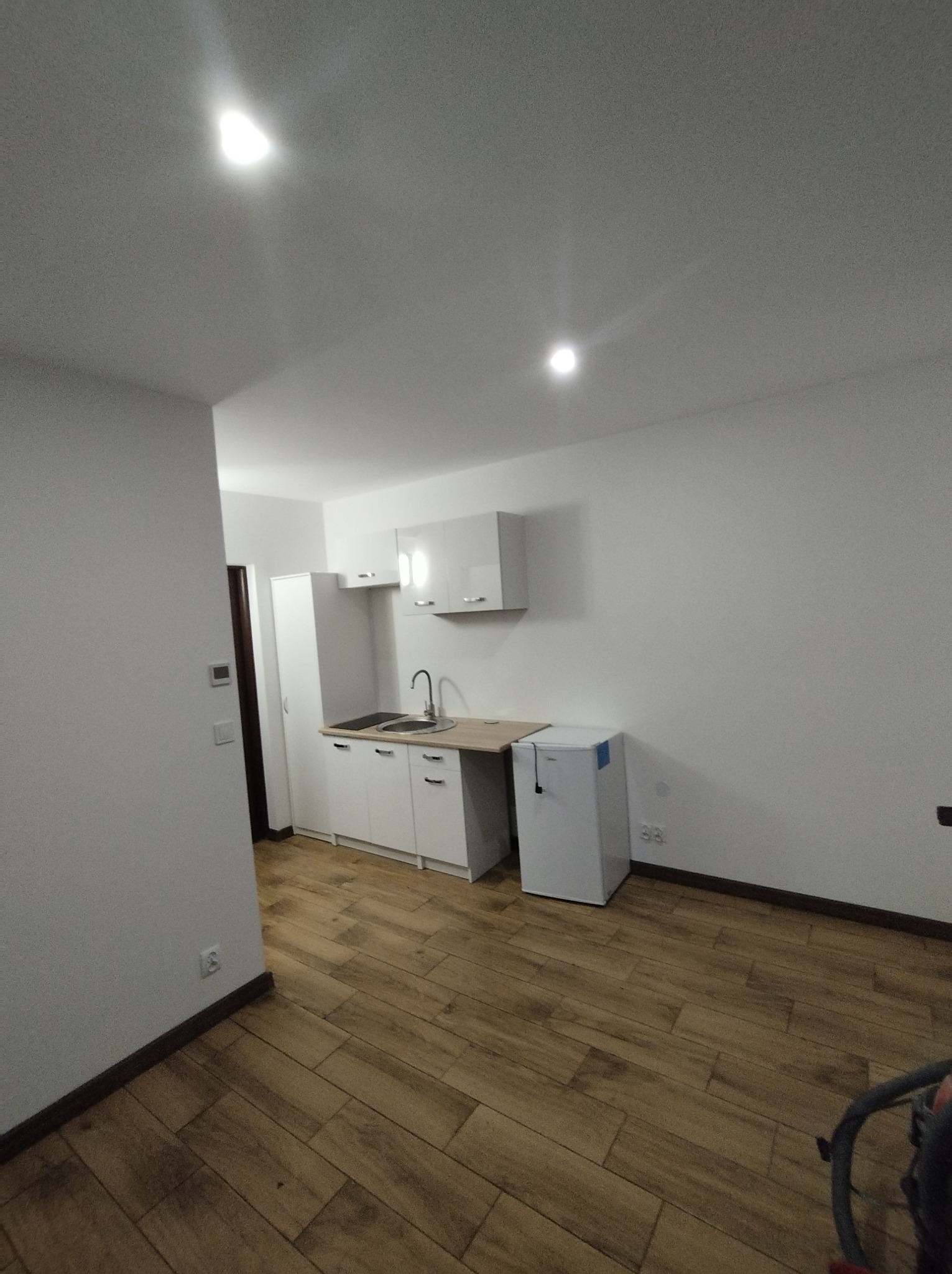 Nowoczesna, minimalistyczna kuchnia w małym mieszkaniu z białymi szafkami, drewnianą podłogą i małą lodówką.