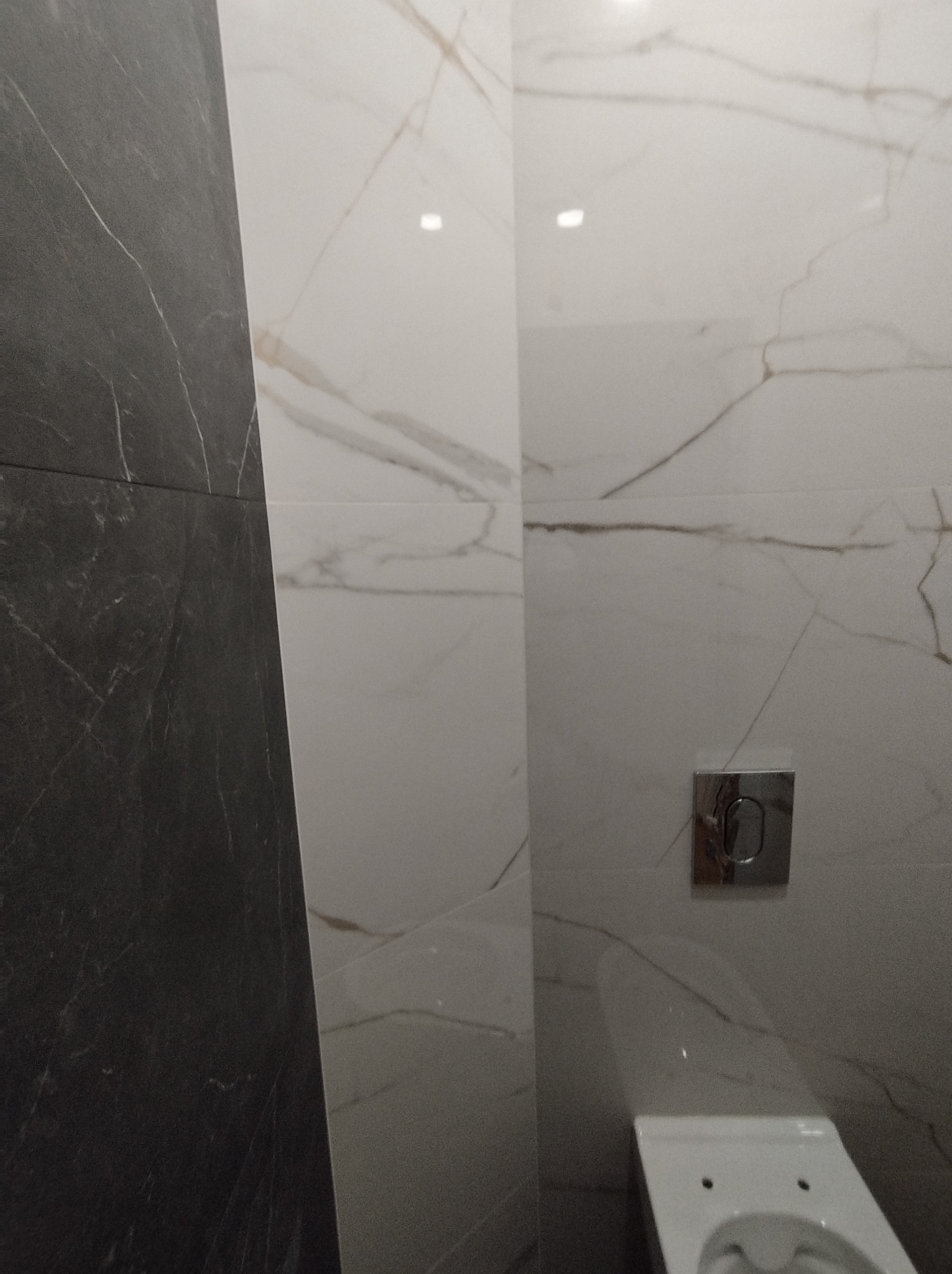 Wykończona łazienka z czarno-białymi płytkami imitującymi marmur, widoczny fragment toalety i przycisk spłuczki.