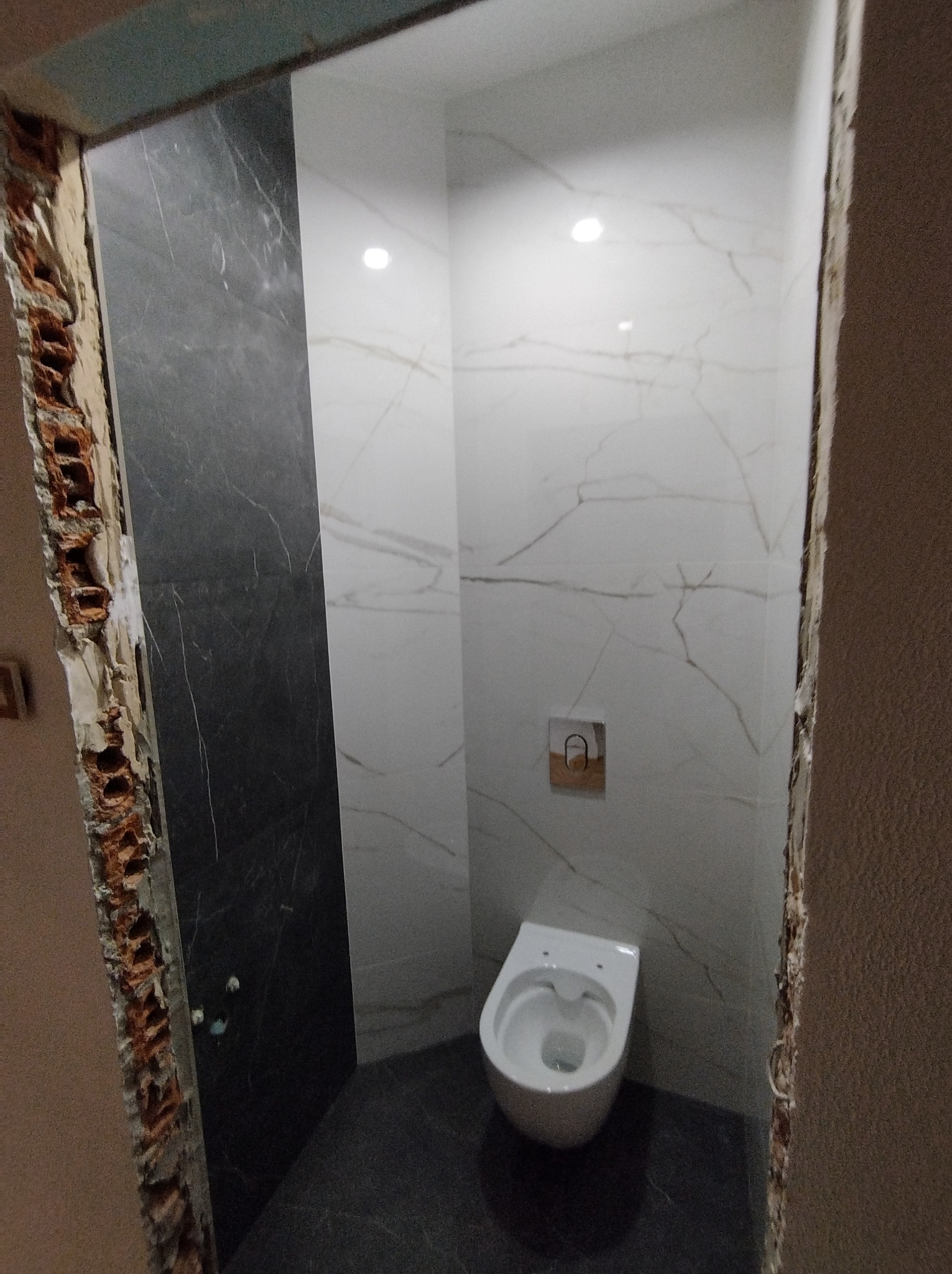 Wnętrze toalety w trakcie remontu: częściowo wykończone ściany z białych i czarnych płytek imitujących marmur, widoczny stelaż podtynkowy i wisząca muszla klozetowa; surowe krawędzie muru.