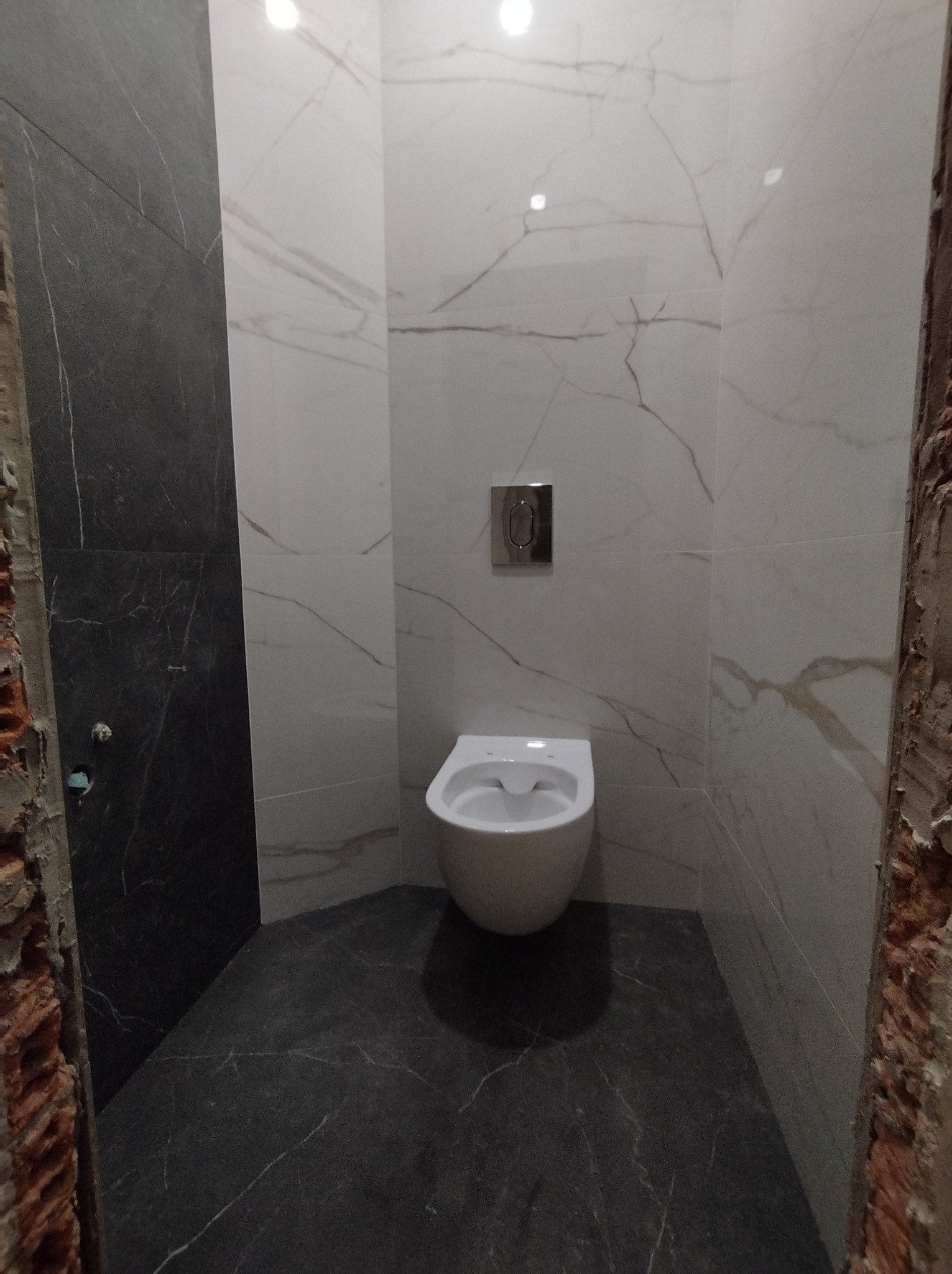 Nowoczesna toaleta podwieszana w łazience wykończonej płytkami imitującymi marmur, z widocznymi surowymi fragmentami muru w trakcie remontu.