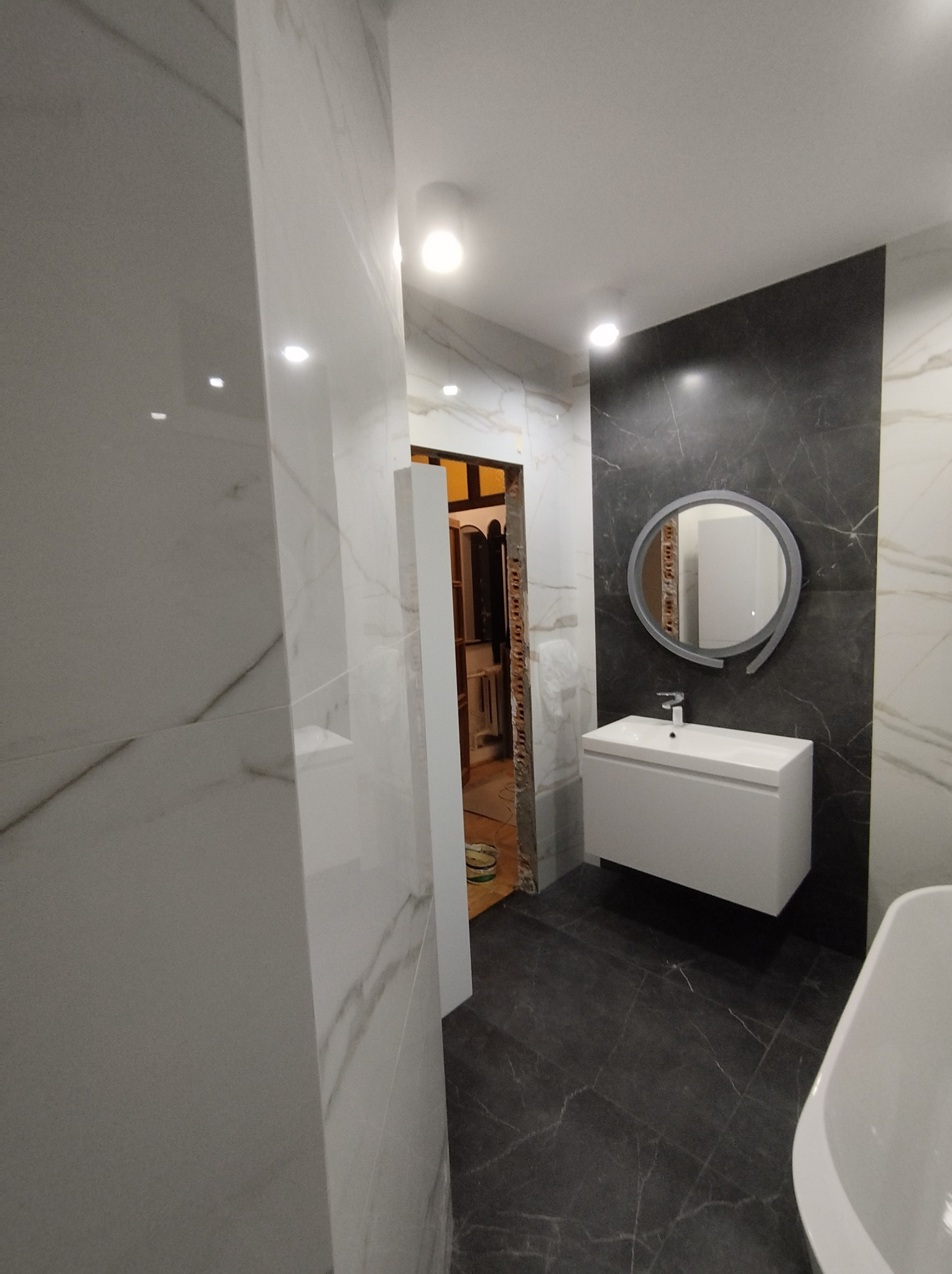 Nowoczesna łazienka w trakcie wykańczania: białe płytki imitujące marmur na ścianach, ciemnoszara podłoga z wzorem żyłkowania, biała umywalka z szafką, okrągłe lustro z podświetleniem, widoczne...