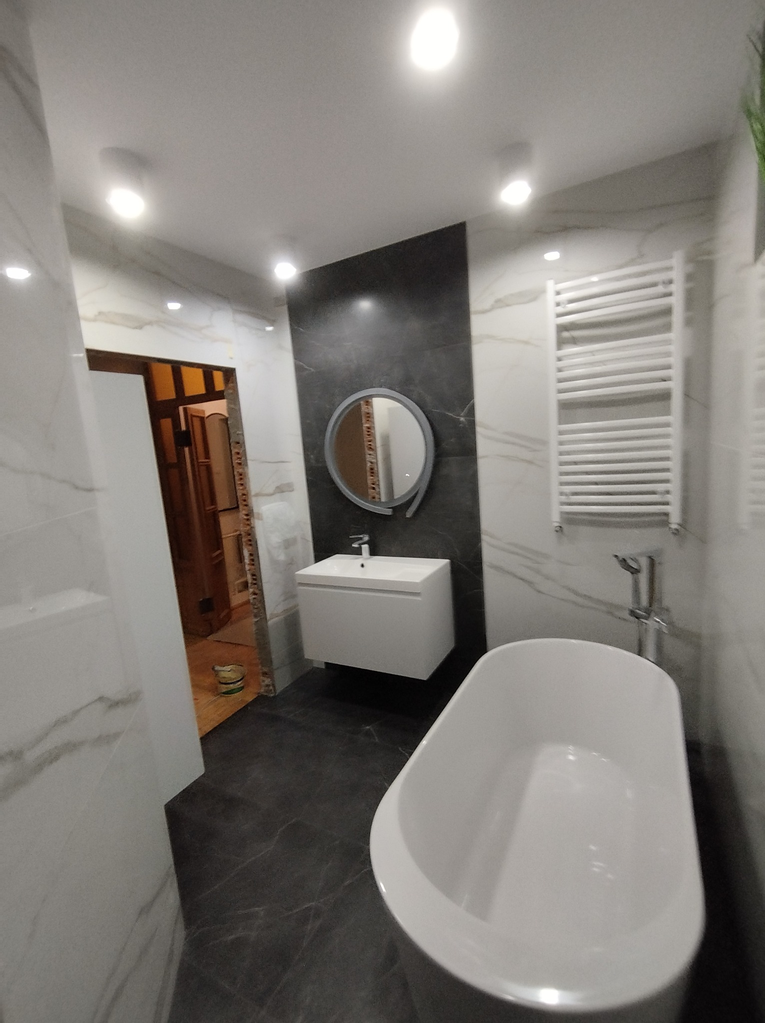 Nowoczesna łazienka z wolnostojącą wanną, umywalką na szafce i grzejnikiem drabinkowym, wykończona płytkami imitującymi marmur i ciemnoszarym akcentem na ścianie, z widocznym fragmentem...