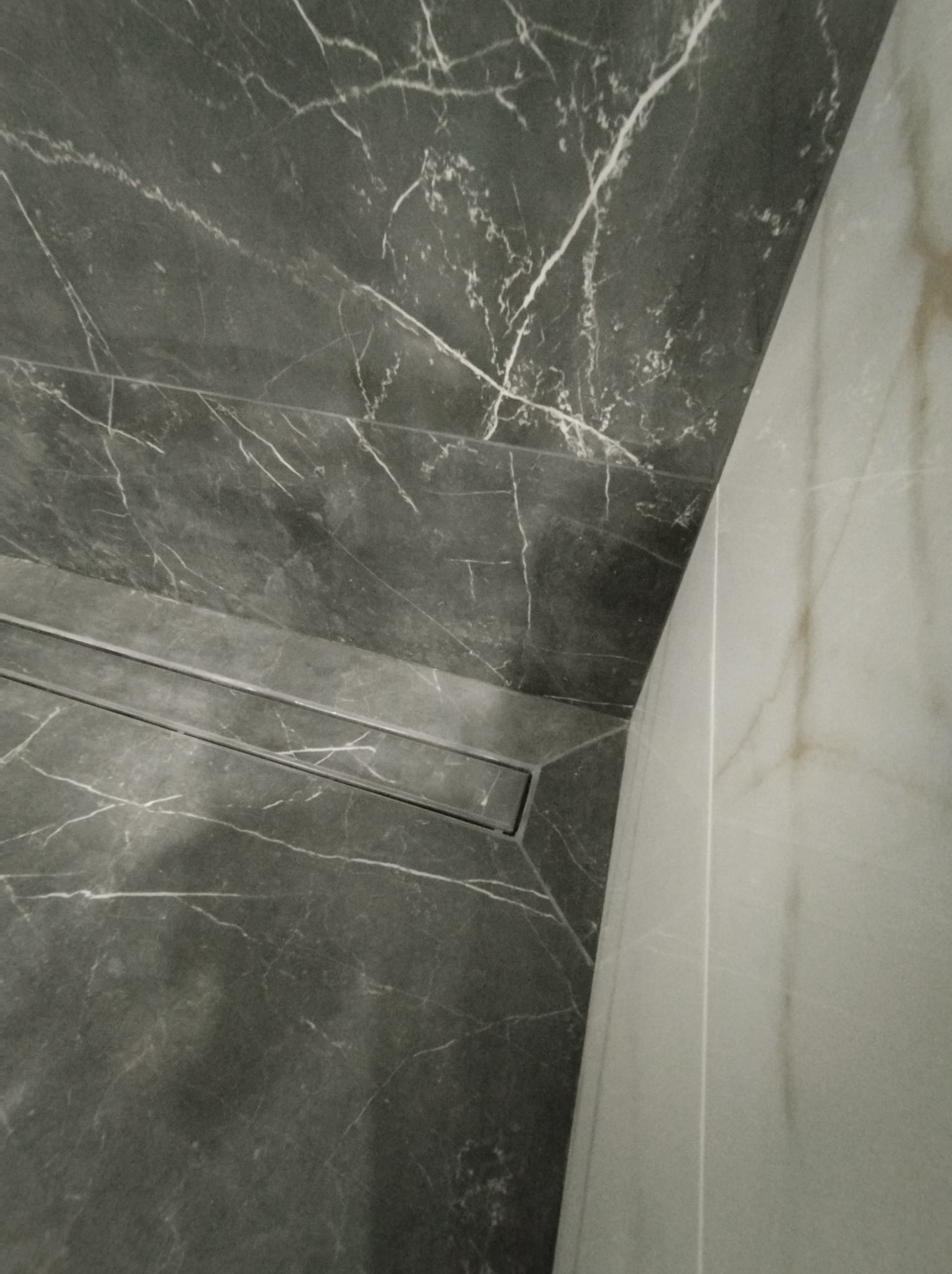 Wykończona łazienka z szarymi płytkami imitującymi marmur z wyraźnym białym żyłkowaniem, widoczny liniowy odpływ podłogowy i fragment jasnej ściany.