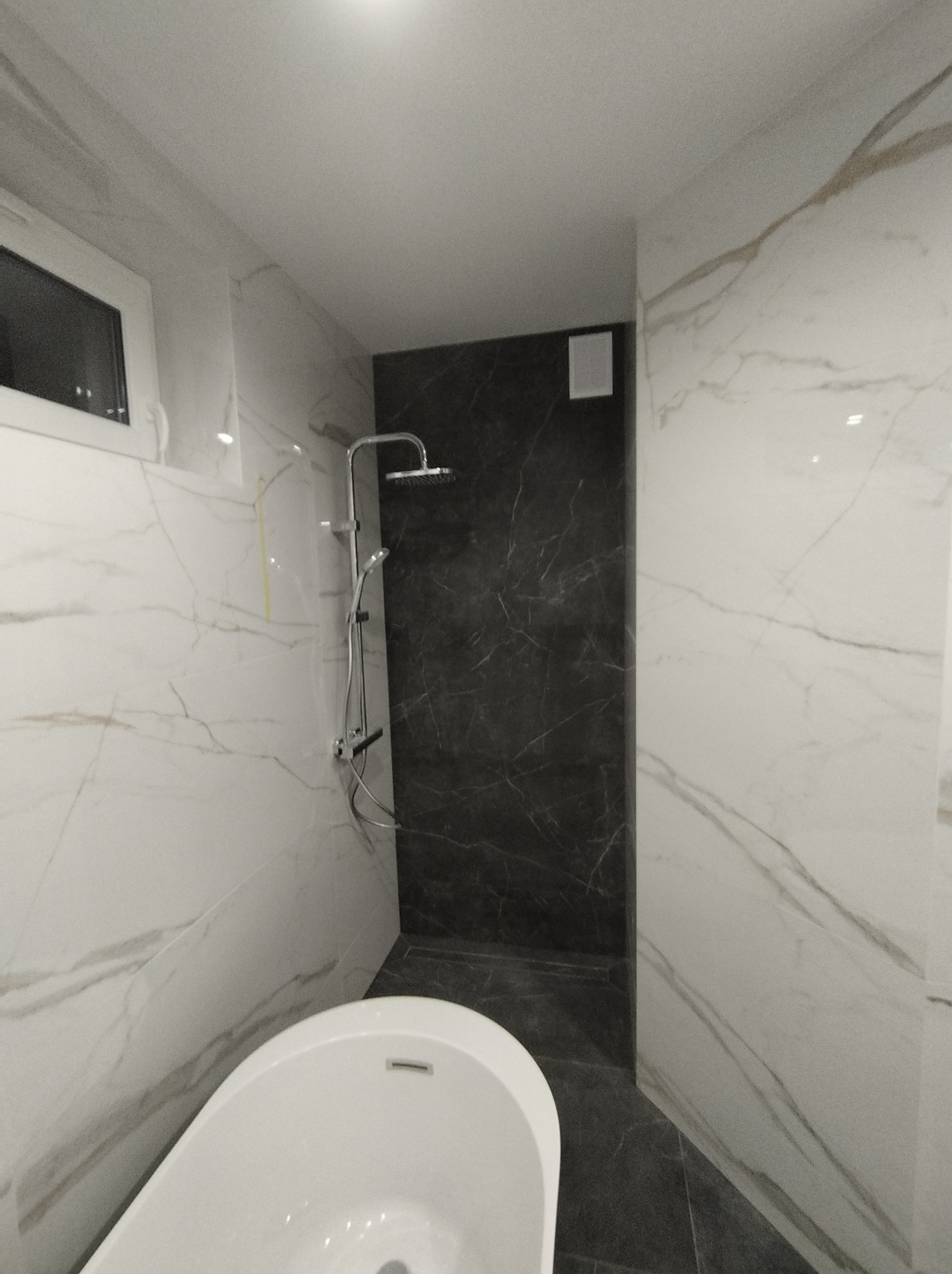 Nowoczesna łazienka z wanną wolnostojącą i kabiną prysznicową wykończona białymi i grafitowymi płytkami imitującymi marmur.