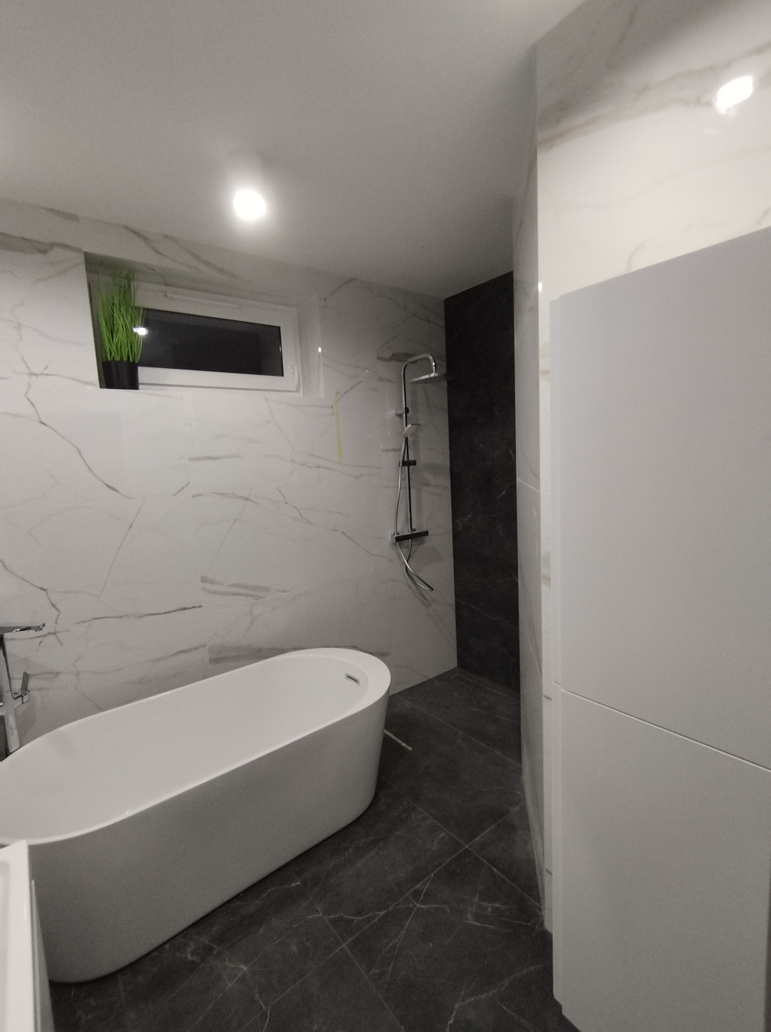 Nowoczesna łazienka z wolnostojącą wanną, marmurowymi płytkami na ścianach i ciemną podłogą, uzupełniona o minimalistyczną armaturę i dekoracyjną roślinę w oknie.