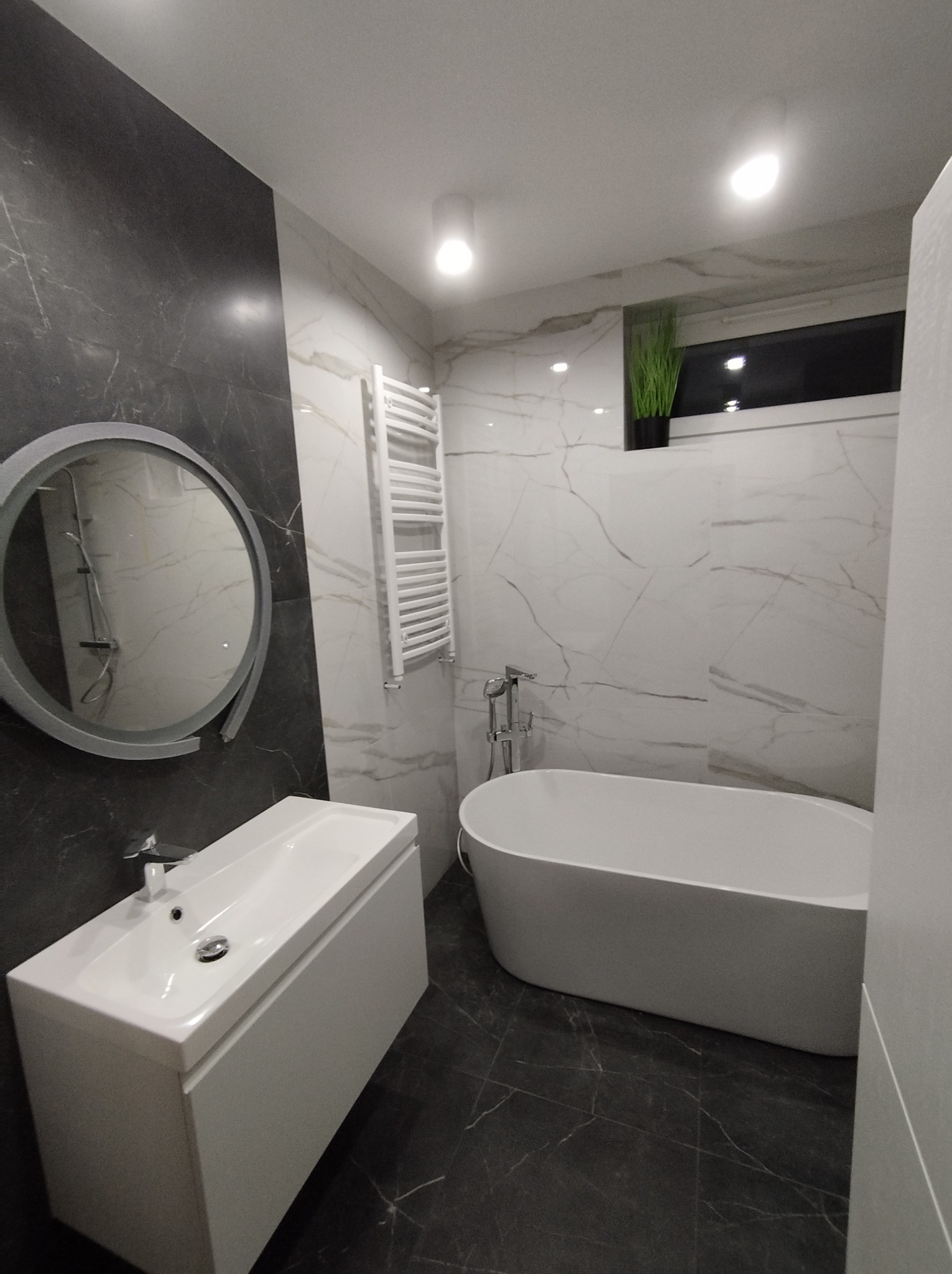Nowoczesna łazienka z czarno-białymi marmurkowymi płytkami, owalną wanną, białą szafką z umywalką i okrągłym lustrem z podświetleniem.