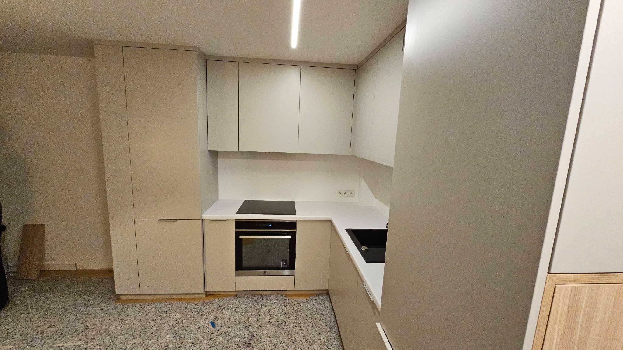 Nowoczesna, beżowa kuchnia z matowymi frontami, czarną płytą indukcyjną i zlewozmywakiem, oświetleniem LED podszafkowym i szarym dywanem na podłodze.