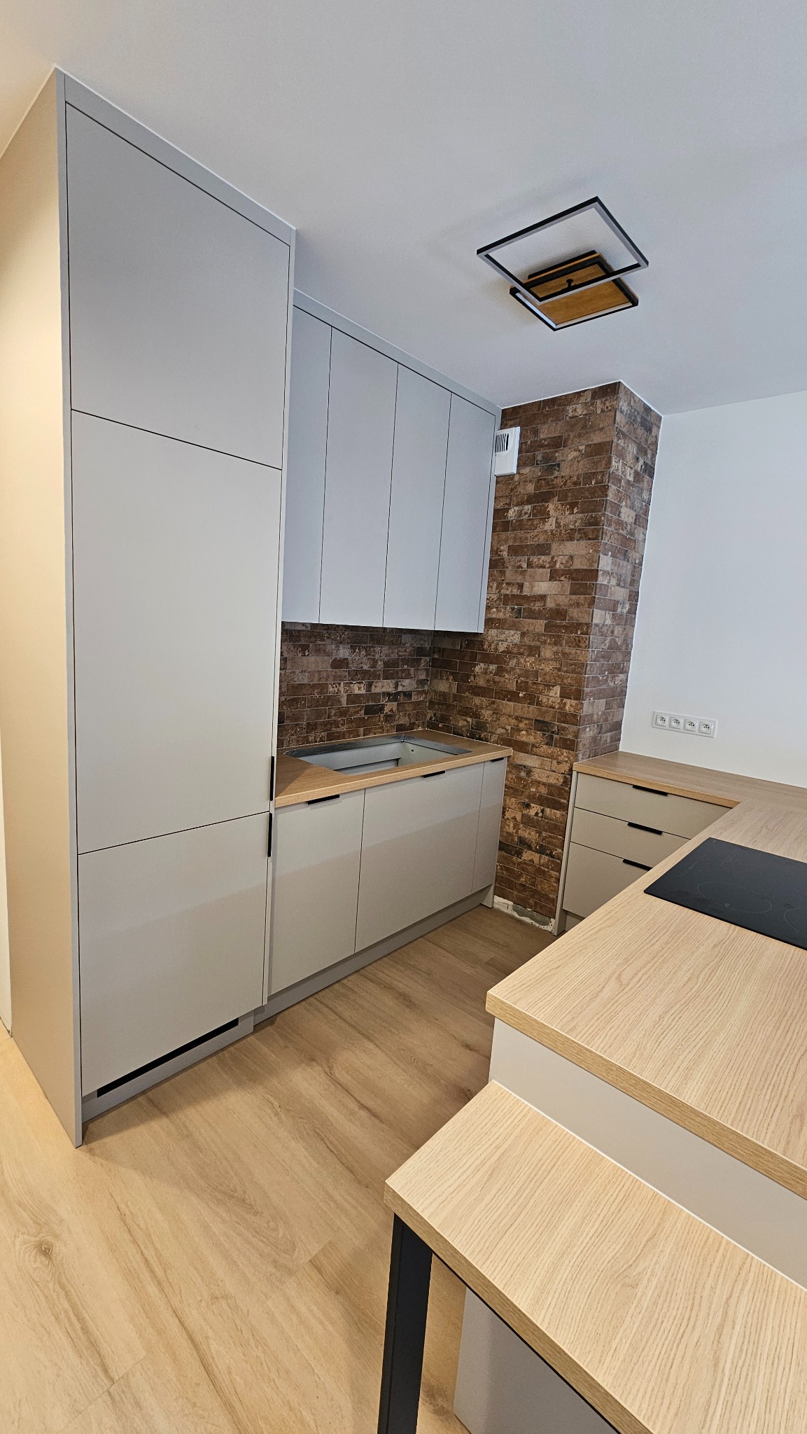 Nowoczesna kuchnia w odcieniach szarości z ceglaną ścianą, minimalistyczne szafki bez uchwytów, drewniany blat i podłoga, oświetlenie sufitowe w geometrycznym stylu.