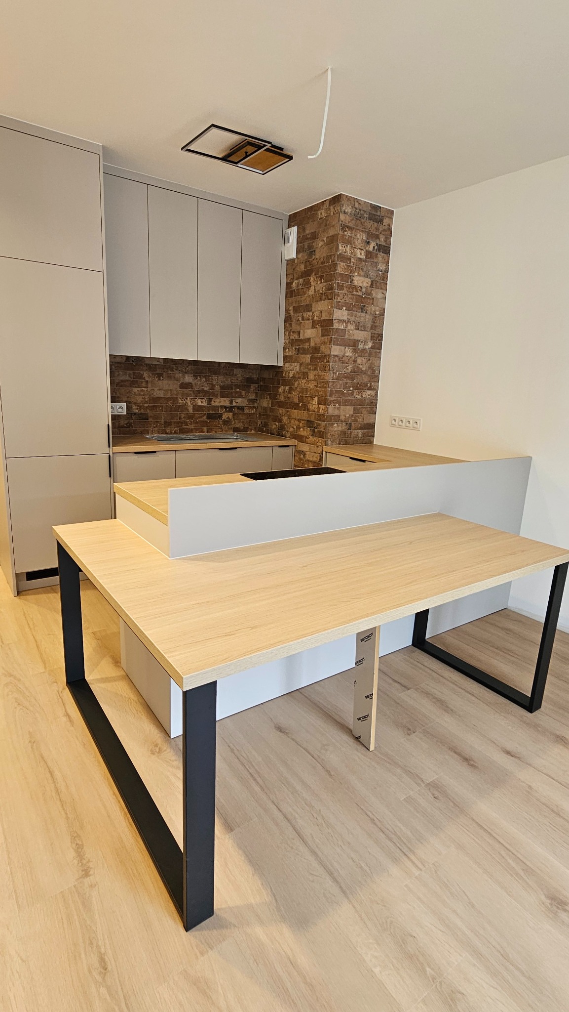 Nowoczesna kuchnia z wyspą i ceglaną ścianą, jasne fronty szafek i blat z drewnopodobnym dekorem, czarne nogi wyspy.