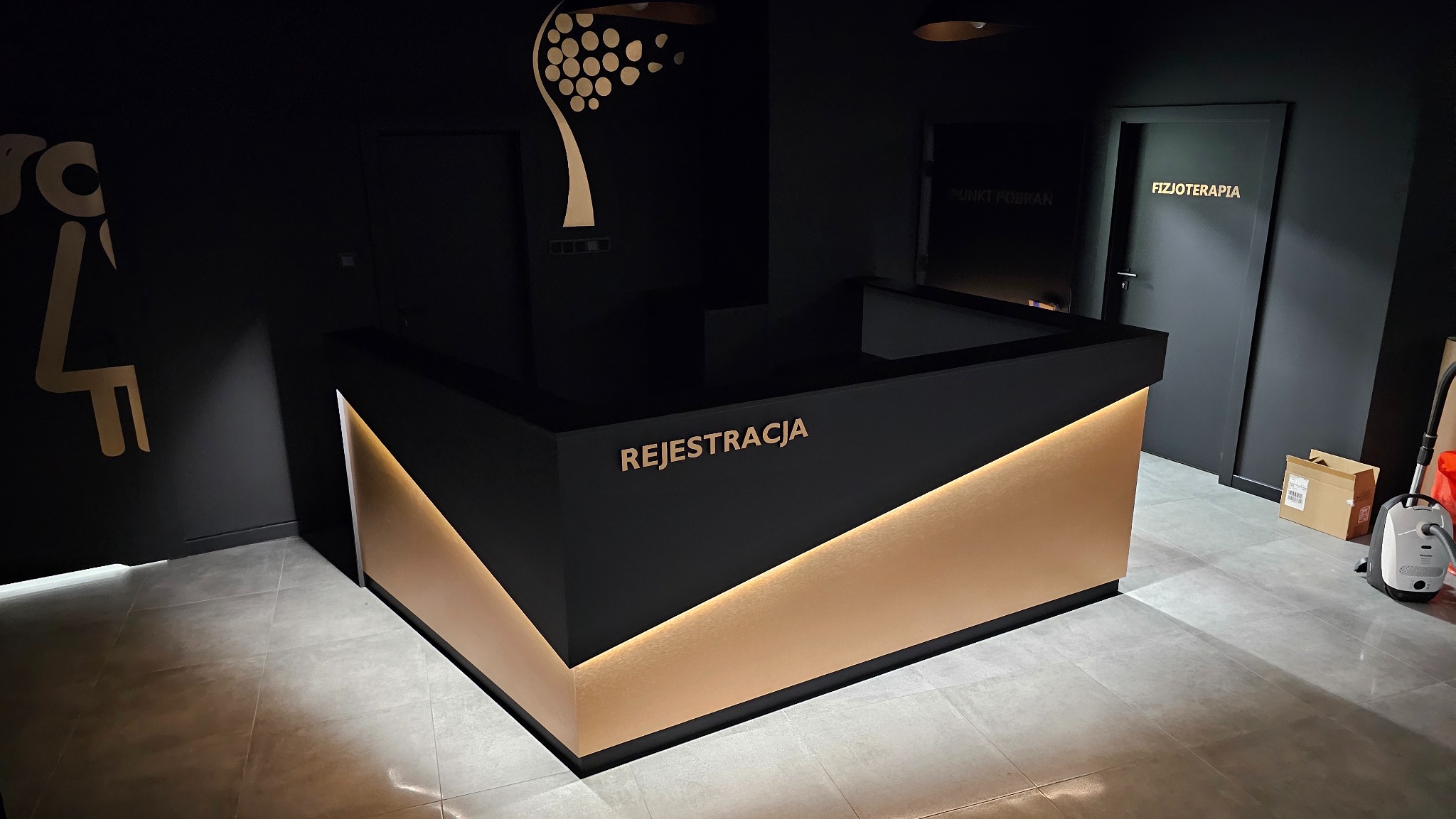 Nowoczesna recepcja z czarnym blatem i złotymi panelami, napis REJESTRACJA, widoczne drzwi z napisem FIZJOTERAPIA i PUNKT POBRAŃ, minimalistyczny design