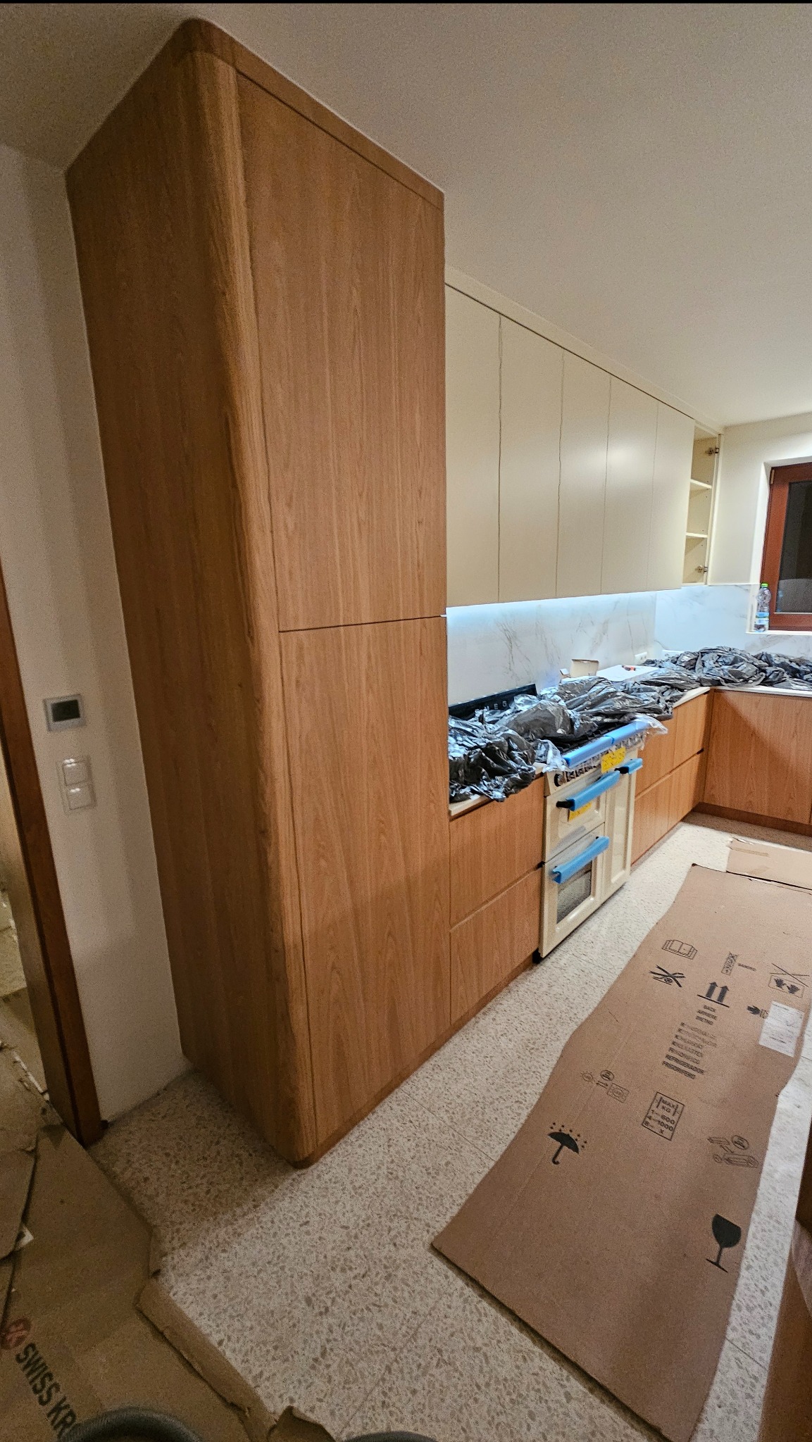 Wysoka, drewniana szafka kuchenna z zaokrąglonymi krawędziami, część nowej zabudowy kuchennej w trakcie montażu, zabezpieczona folią i kartonami.