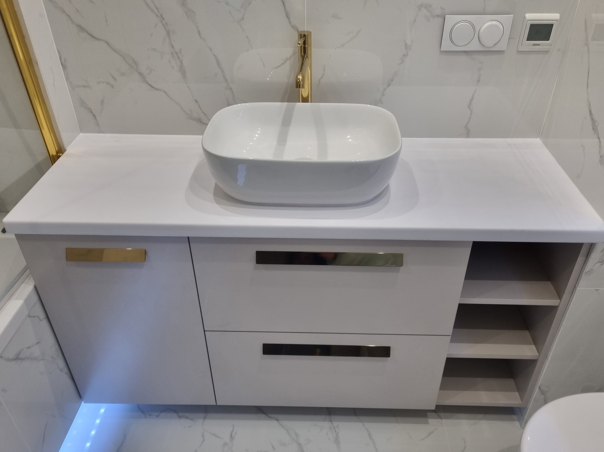 Nowoczesna łazienka z białą, nablatową umywalką o zaokrąglonych kształtach, złotą baterią i szafką z szufladami oraz półkami, podświetlana od dołu.